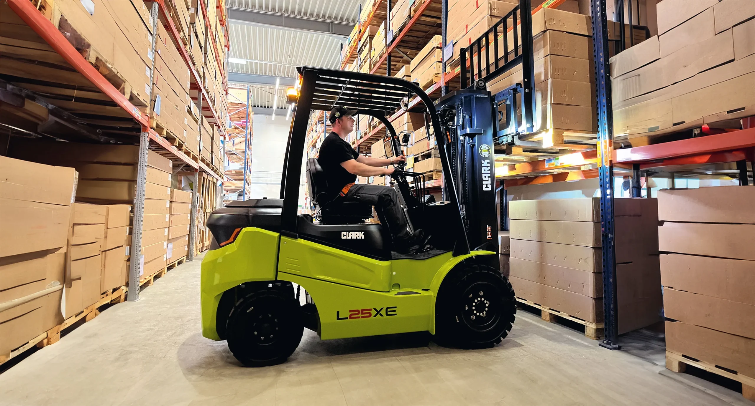 La carretilla eléctrica CLARK L25XE maniobrando entre estanterías de un almacén mientras eleva una carga de pallets.