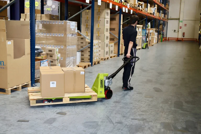 Trabajador utilizando una transpaleta manual verde para transportar cajas en un almacén con estanterías llenas de mercancías.