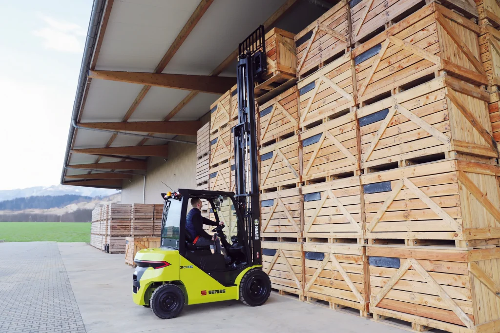 Carretilla elevadora eléctrica CLARK S30XE levantando una caja de madera en un almacén al aire libre, con un operario manejándola.