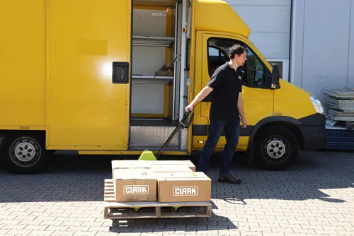 Trabajador utilizando una transpaleta manual CLARK para mover un palet con cajas marcadas con el logo CLARK frente a una furgoneta amarilla.