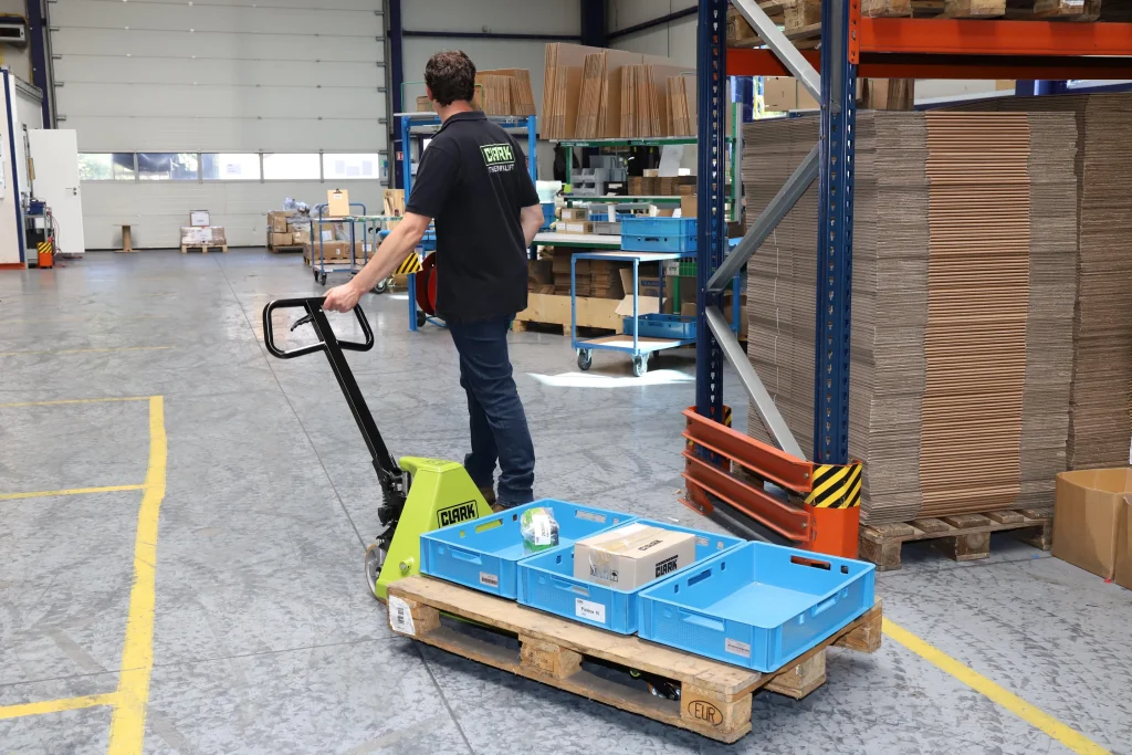 Operario manejando una transpaleta manual CLARK para mover un palet con cajas plásticas en un almacén organizado.