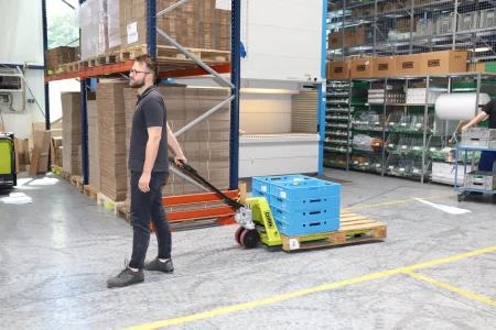 Trabajador manejando una transpaleta manual con cajas de plástico azul sobre un palet en un almacén bien organizado.